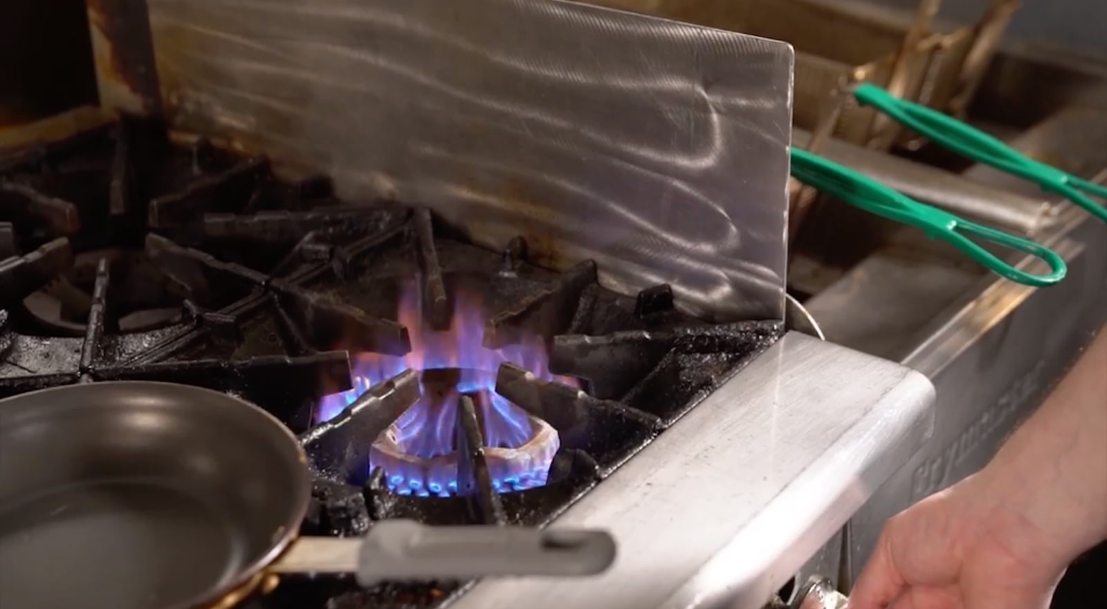 A hand turns up the heat on a gas stove. A non-stick pan sits to the left of the flame.
