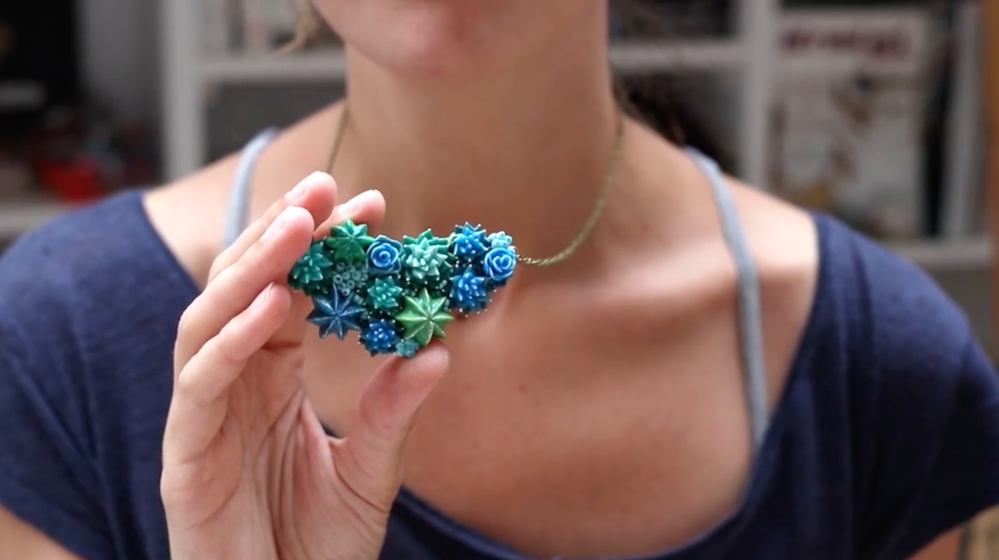 A woman in a navy blue shirt holds a blue and green succulent necklace sculpted from polymer clay.