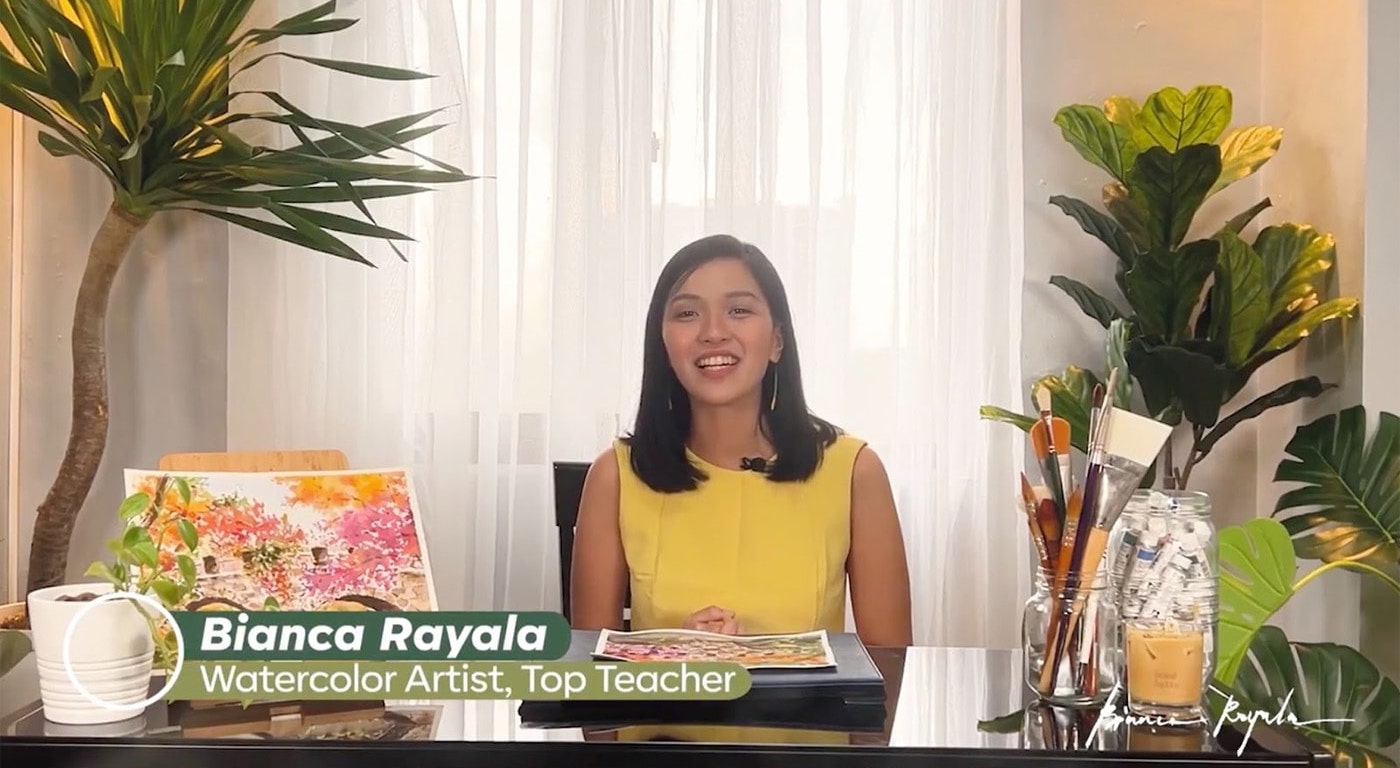 Teacher Bianca Rayala sits at her desk. One of her watercolor landscapes is displayed to her left, and several large plants sit behind her. You can also see a jar of her paintbrushes, and a jar of paints. 
