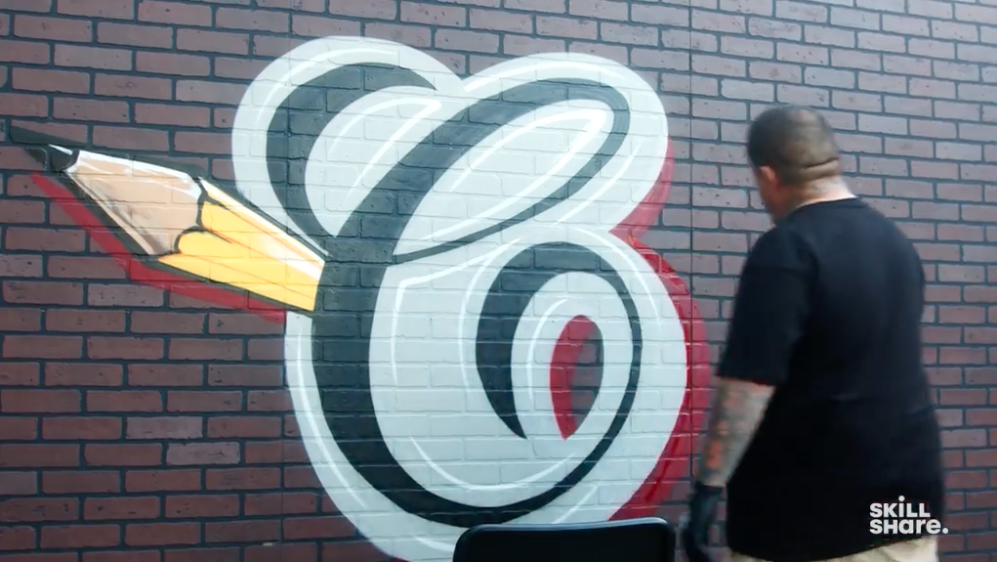 A man stands in front of a large brick wall, where he has just painted a logo featuring a cursive "C" with a pencil coming out of one side. The man is dressed in all black and has several visible tattoos.