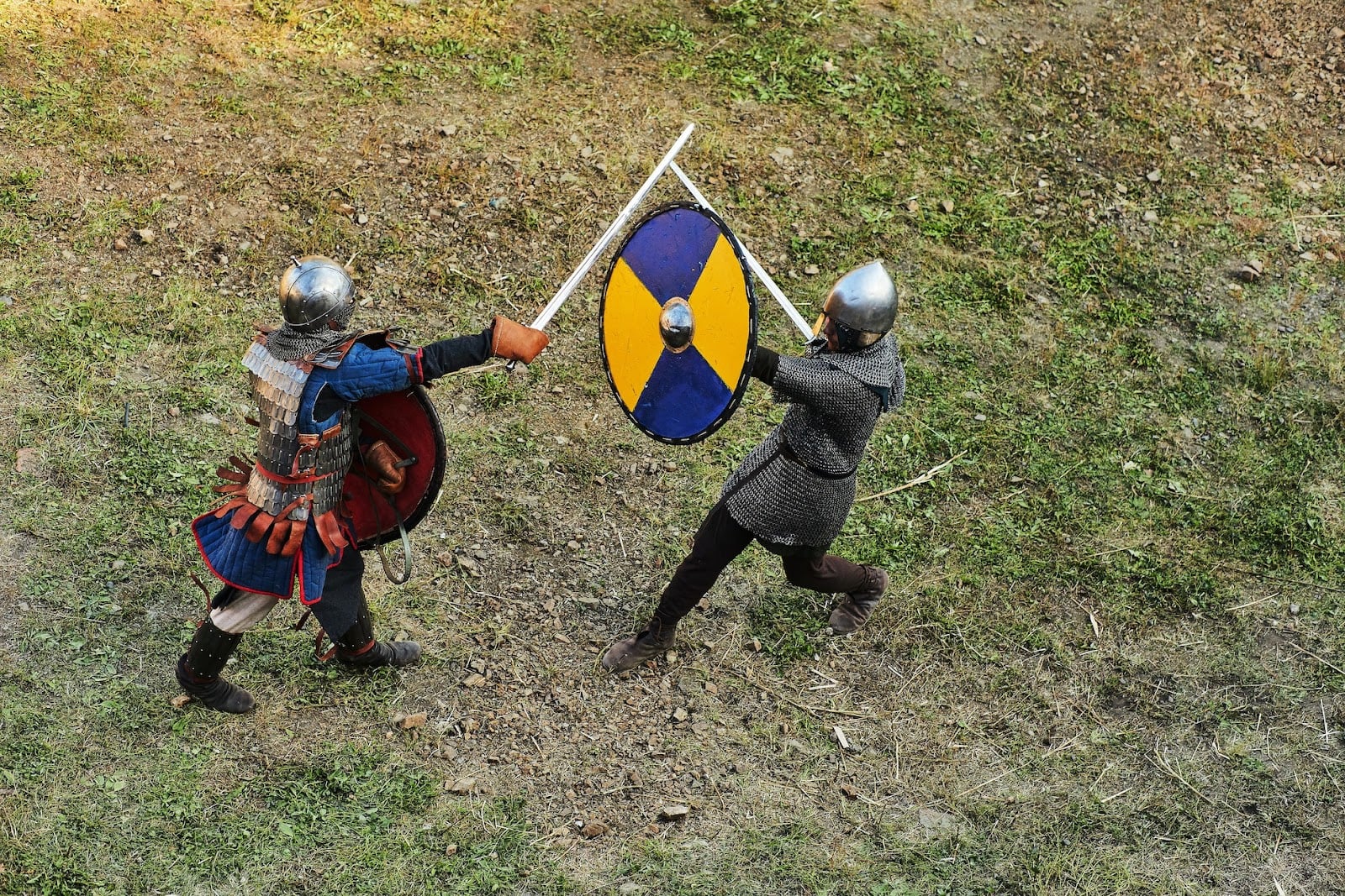 Two people on a grass field fight with swords and shields. Both are wearing metal helmets, and one is wearing a chain mail suit, while the other dons a medieval armor vest.