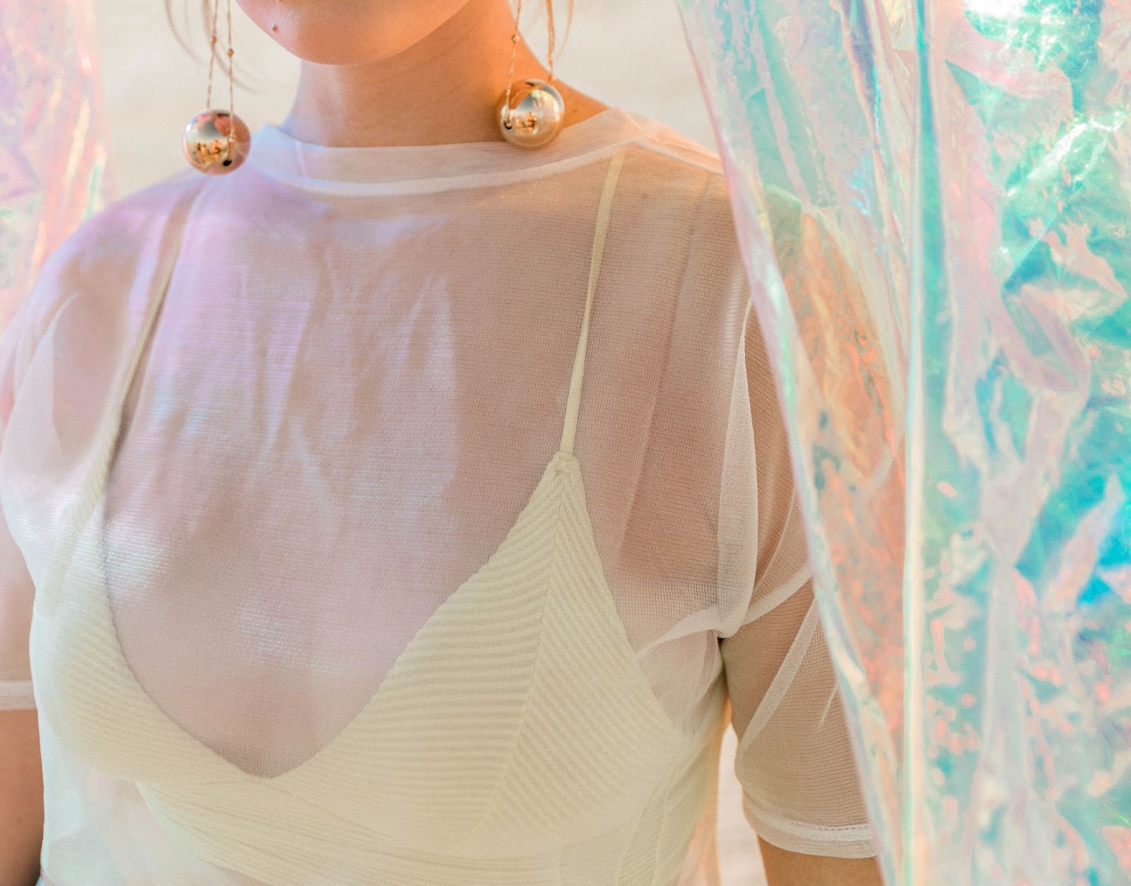 A woman’s torso featuring her long statement earrings with dangling gold orbs and sheer, white blouse.