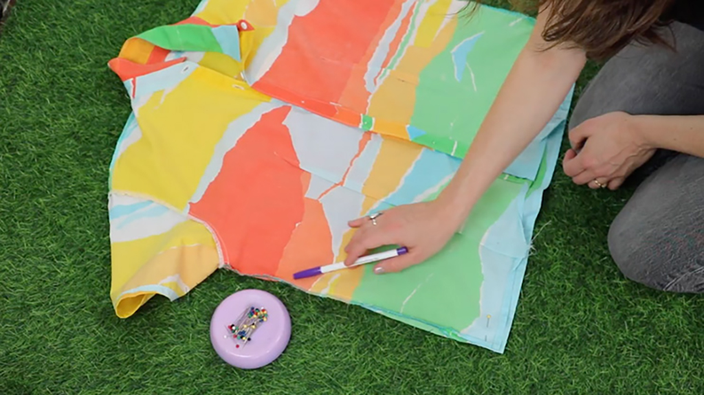 Kneeling in the grass, the artist points to the edge of a button up shirt with a purple marker. The button up shirt is red, yellow, orange, green, and blue. It also lays in the grass next to some sewing pins.