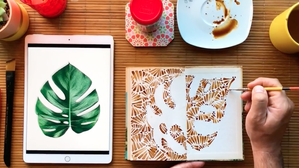 On a bamboo placemat sits a tablet with a painting of a large green monstera leaf. Next to it is an open notebook, with a hand panting the outline of a monstera in various shades of brown. At the top of the frame sits a cup of coffee, and a saucer next to it with coffee spilled out on it.