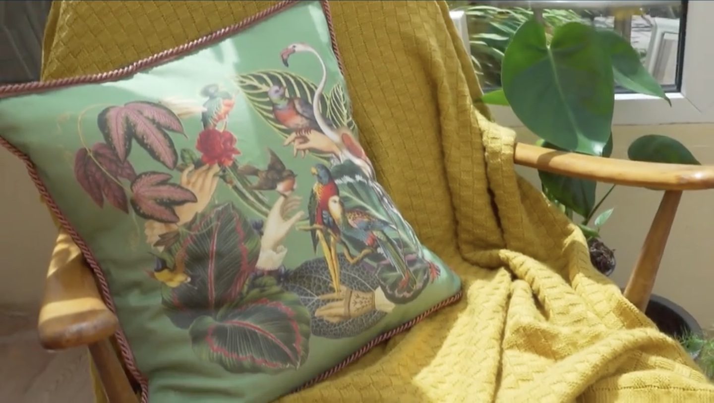 Square pillow with a green background with several types of birds and multicolored leaves, and a braided trim around its perimeter. It sits on a yellow throw that's draped over a wooden chair.