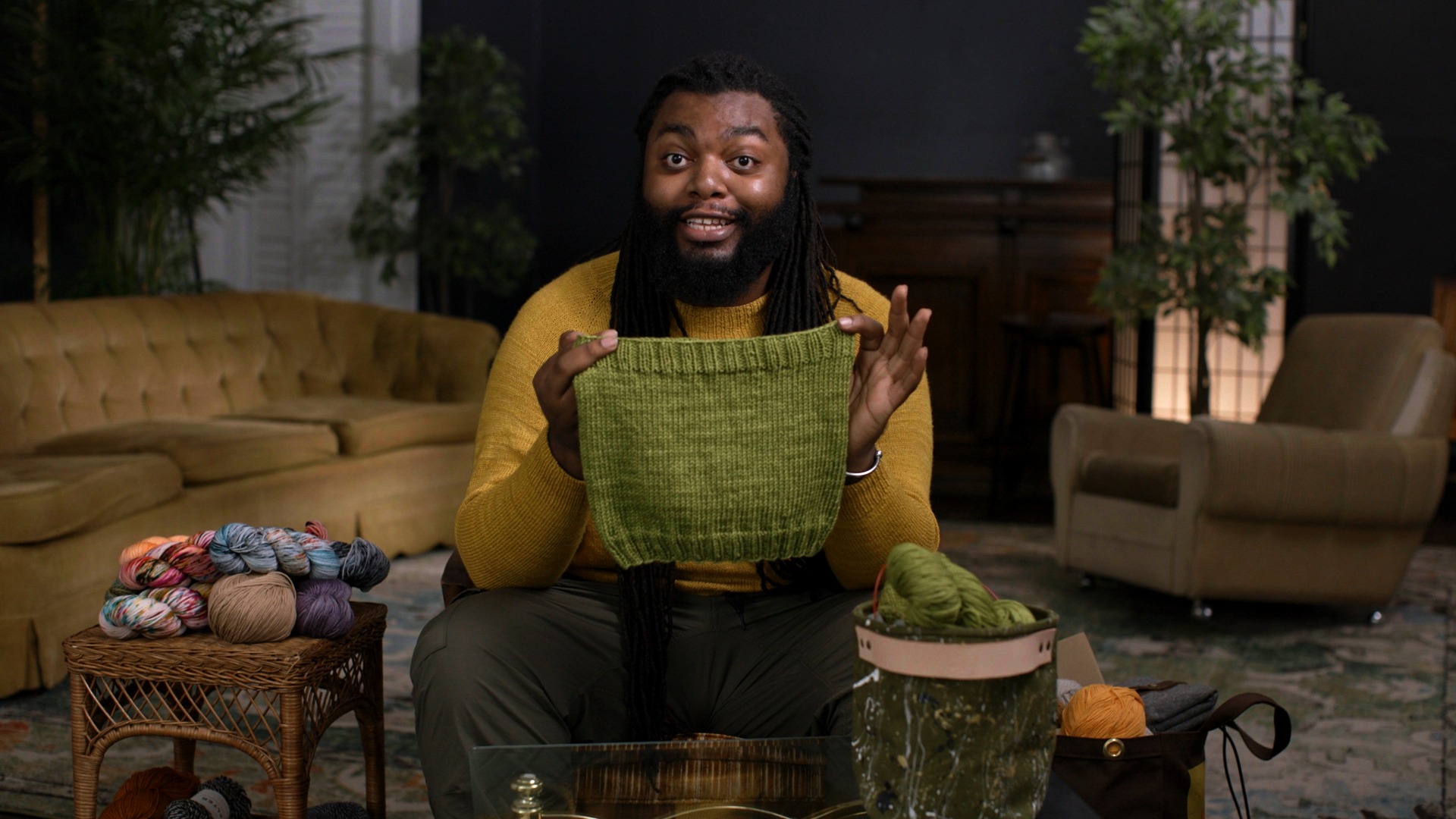 An artist in a yellow sweater sits in a living room holding a green, knitted cowl. To the left a few rolls of yarn sit on a wicker table. The right two bags of yarn are visible.