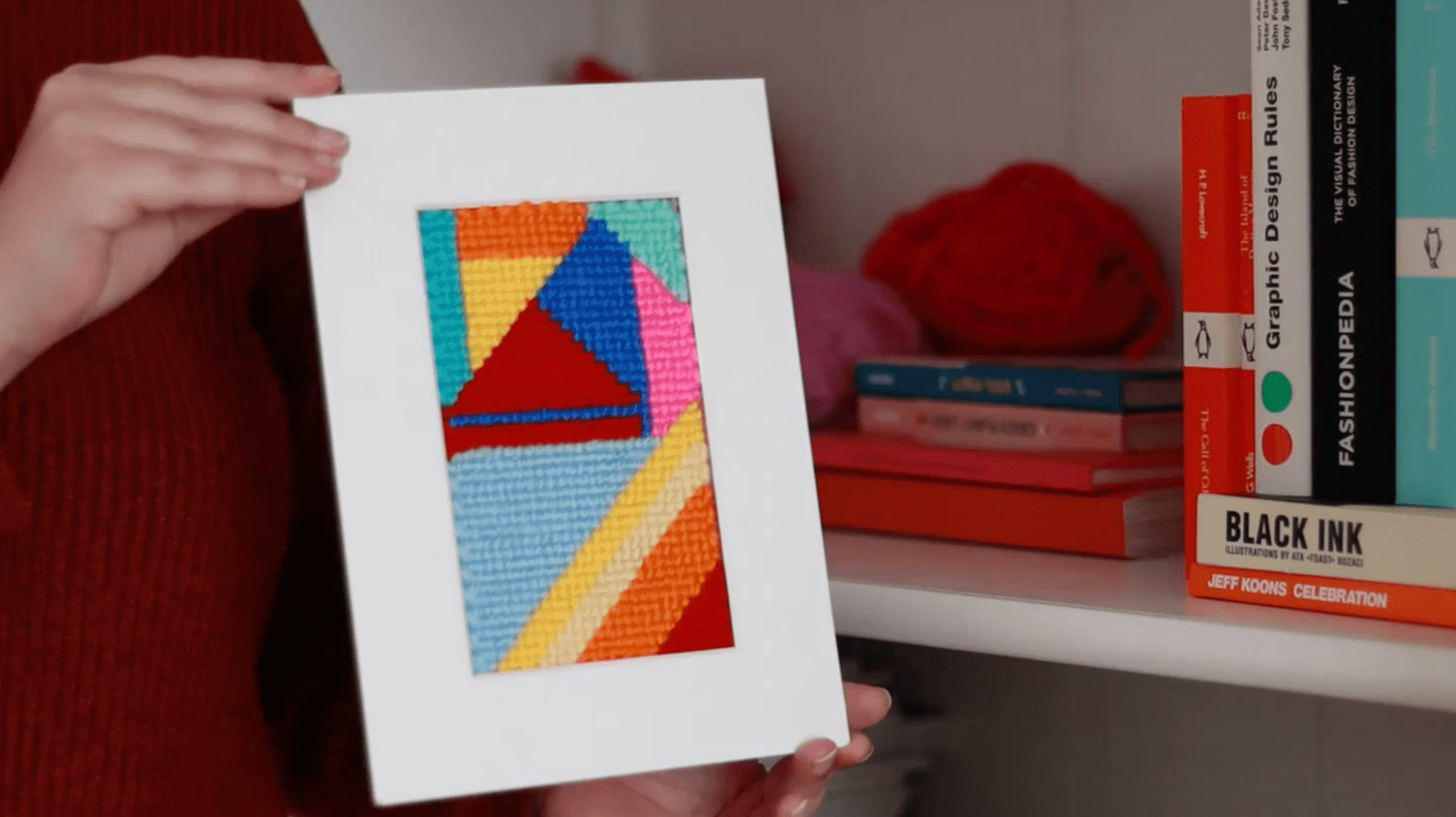 Someone in a red sweater holds a framed needlepoint project. It’s an abstract work made of blues, oranges, pinks, and reds. In the background, a bookshelf with a few books and balls of yarn is visible.