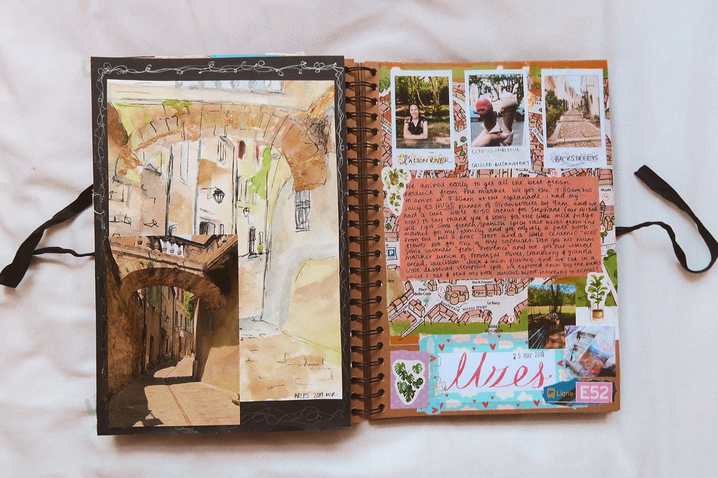 An open scrapbook sits on a white background. On the left page is a photo and a watercolor painting of a stone, pedestrian street. On the right are three polaroid pictures and a variety of stickers, writings, and other paper-based cutouts.