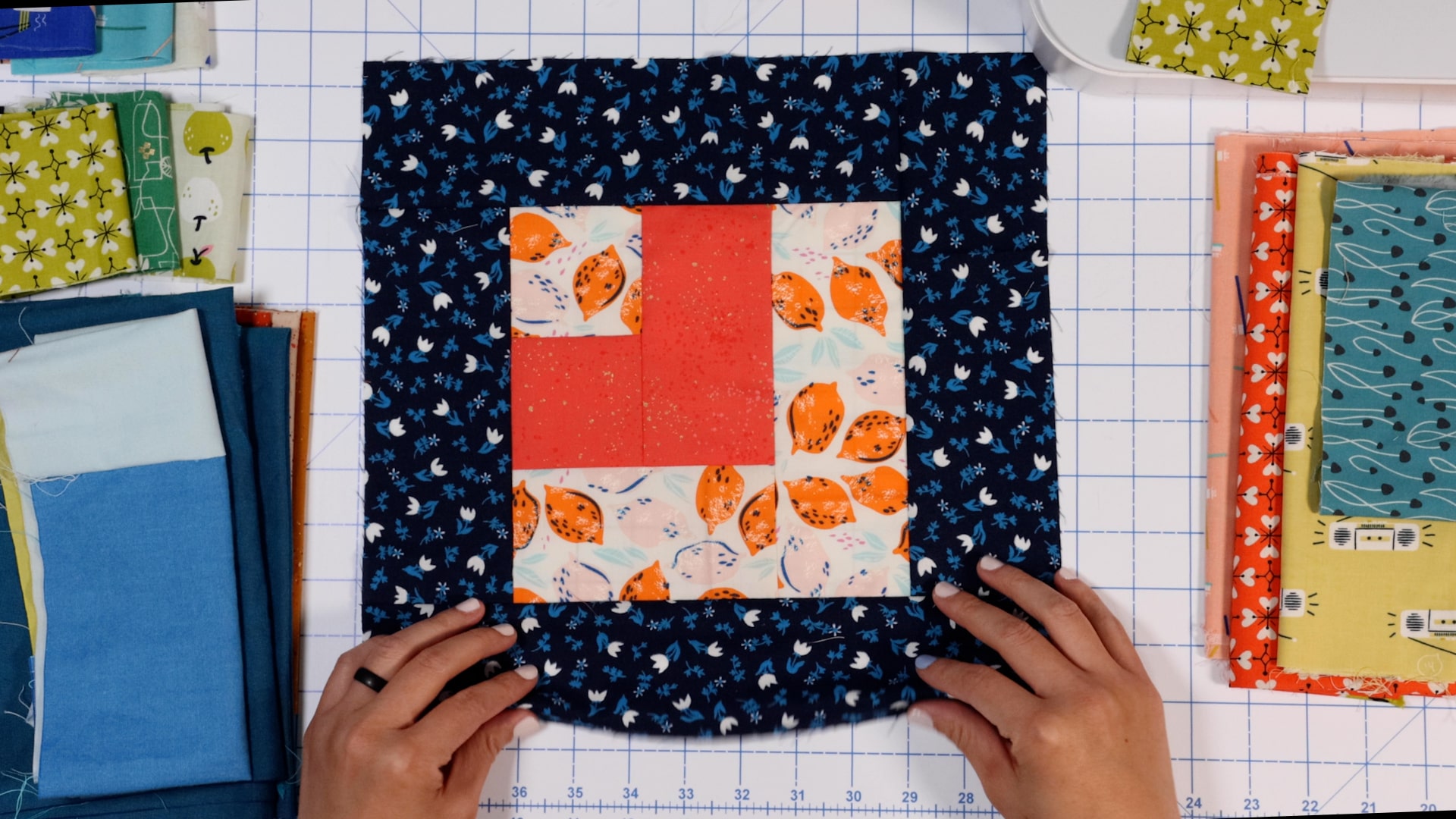 Two hands sit at the bottom left and right edges of a quilted square. Made with blue and white flower fabric, lightly patterned red fabric, and orange and pink lemon fabric, it sits next to a variety of other fabrics.