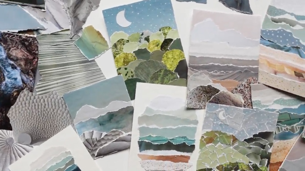 A wide variety of torn paper art collages lay on a white table. Crafted with natural colors like grays, light blues, and greens, an artist has created a variety of scenes including mountains, waves, and the moon.