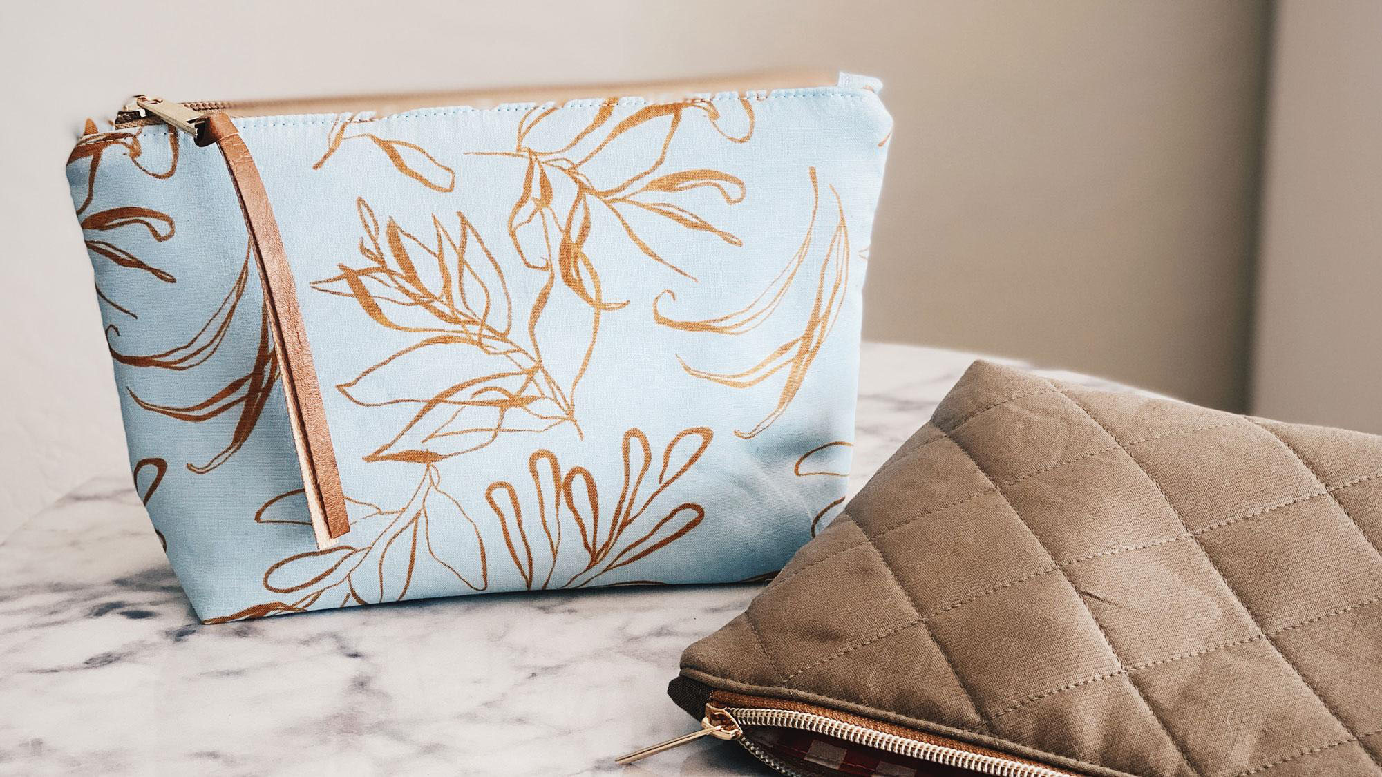A blue, zippered pouch covered in abstract gold leaves sits on a white marble table. A brown, quilted and zippered pouch lies on its side next to the blue pouch.