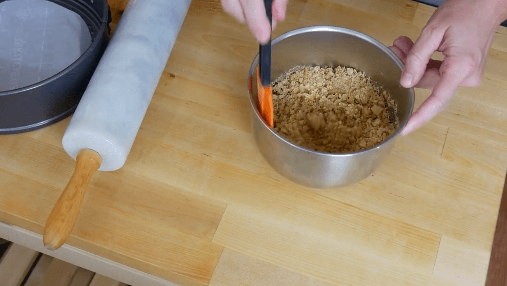 ookie crumbles are mixed in a metal bowl next to a rolling pin, ready to be made into a cheesecake base.