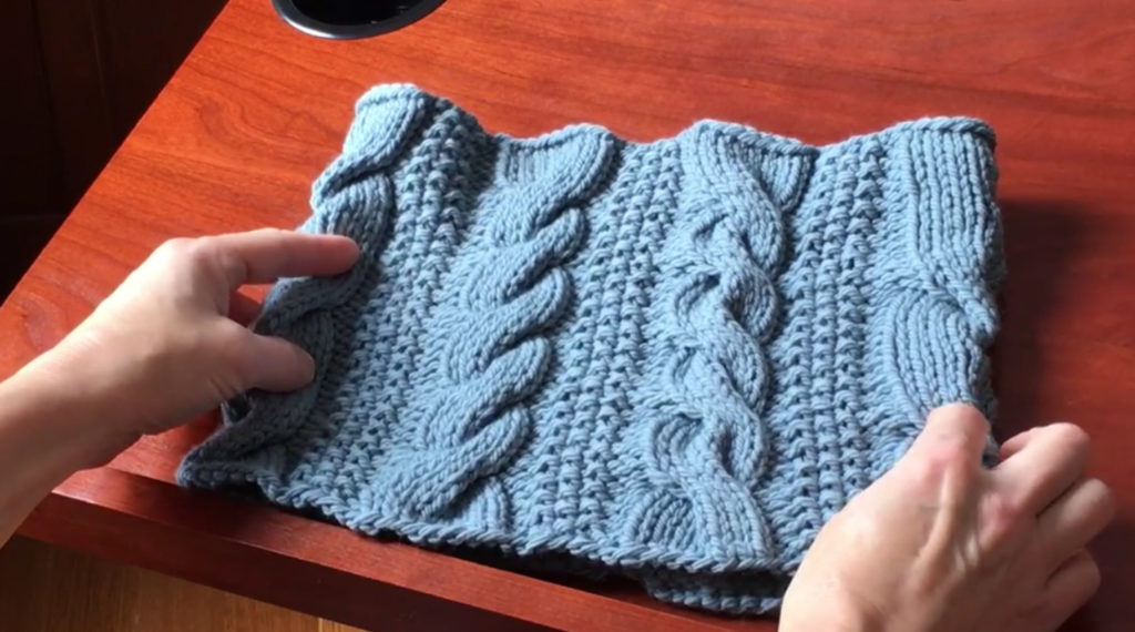 A woman’s hands holding a cowl knit from a soft blue-colored yarn. The cowl has been decorated with several types of knit cables, each one running vertically. 