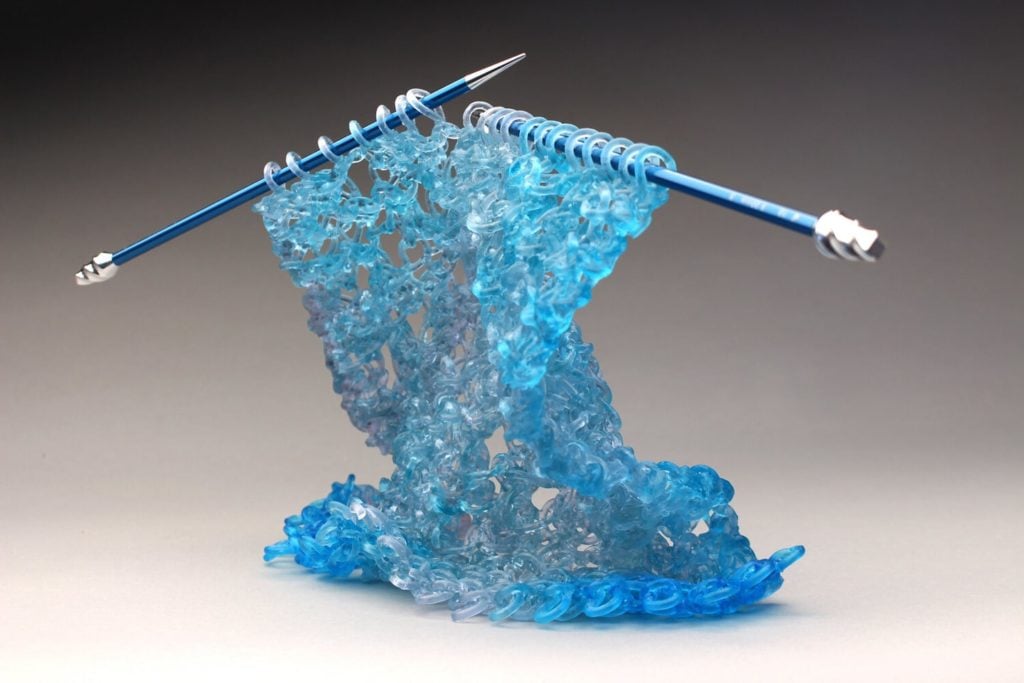 A sculpture of blue knitting needles in the process of knitting blue fabric, with the fabric being made entirely of glass.