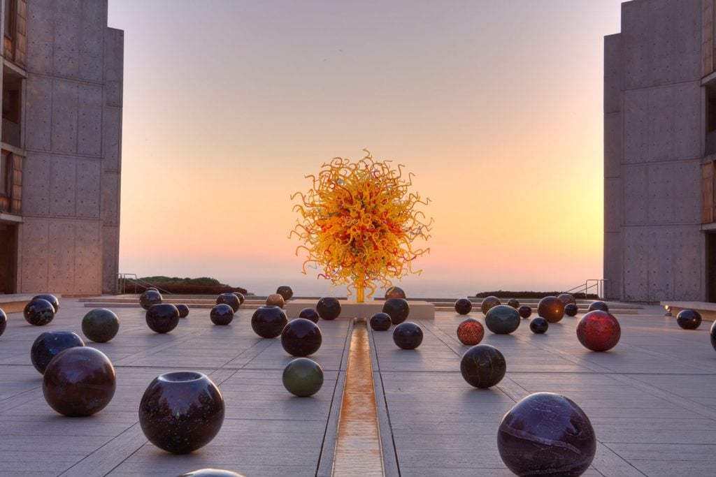 A large sun-like sculpture consisting of hundreds of pieces of spiraling yellow, orange and red glass. On the cement ground surrounding it are dozens of smaller round glass orbs, all in deep shades of red, blue, green and orange.