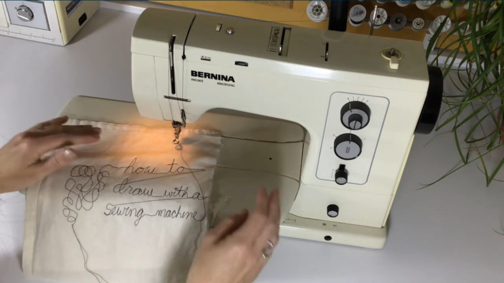 A woman’s hands holding a square of cream fabric on the bed of a sewing machine. The fabric is embroidered with the words ‘how to draw with a sewing machine.’