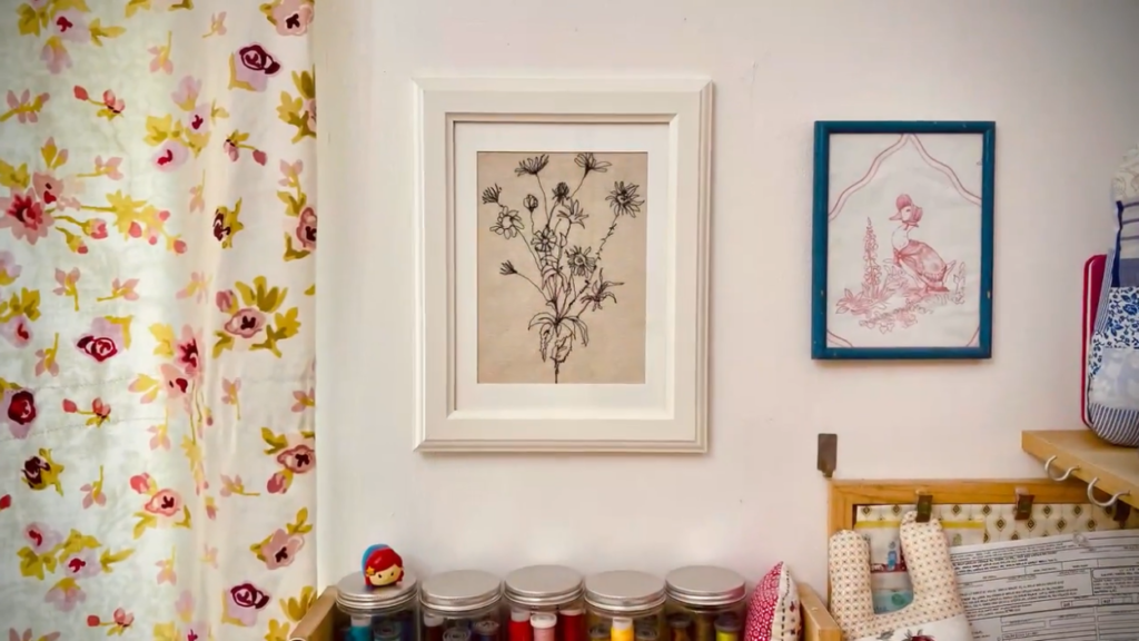 A framed embroidery project hanging on the wall next to curtains and another piece of framed art. It consists of a bundle of wildflowers embroidered in black thread on beige fabric. 