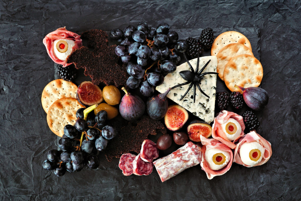 A variety of fruits, meat, cheese, and crackers sit on a dark granite background. A plastic spider and eyeball-inspired mozzarella balls play off the Halloween themed charcuterie board.