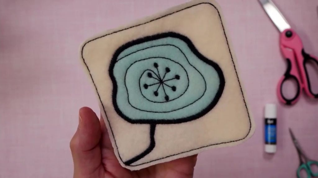 A woman’s hand holding a cream-colored felt coaster over a pink background. The coaster has been embroidered with a simple design of a blue and black flower. 