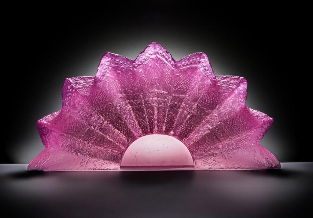A bright pink block of glass which has been sculpted into the shape of a folding fan. The fan is sitting on a black surface in front of a black background, and its glass surface is textured with intricate dotted designs.