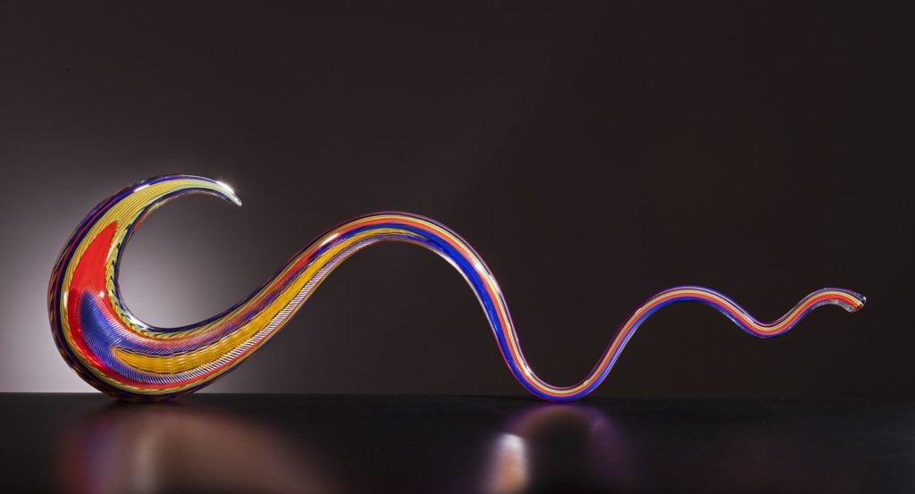 A brightly multi-colored glass sculpture in the shape of a tapered spiral, set atop a black surface in front of a dark gray background.