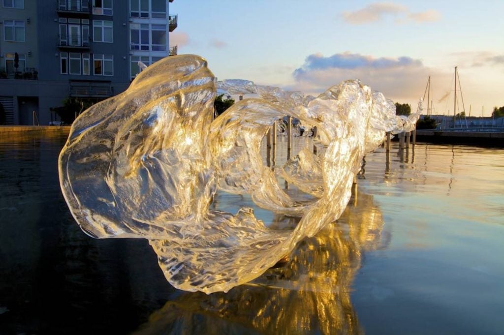 A large, clear glass sculpture in an undulating, abstract shape appears to hover on the surface of the water just outside a large waterfront building. In the background, sailboats float on the water underneath a sunset.