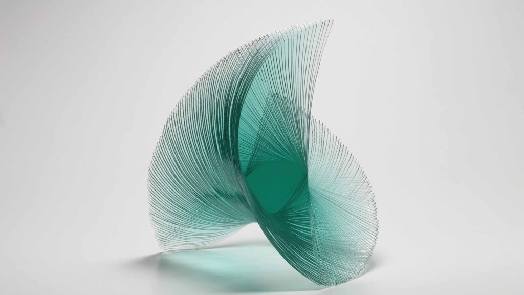 Layered sheets of glass formed into an abstract geometric shape. The sculpture has a blue-green tint, while the surface it rests on and the background behind it are white.