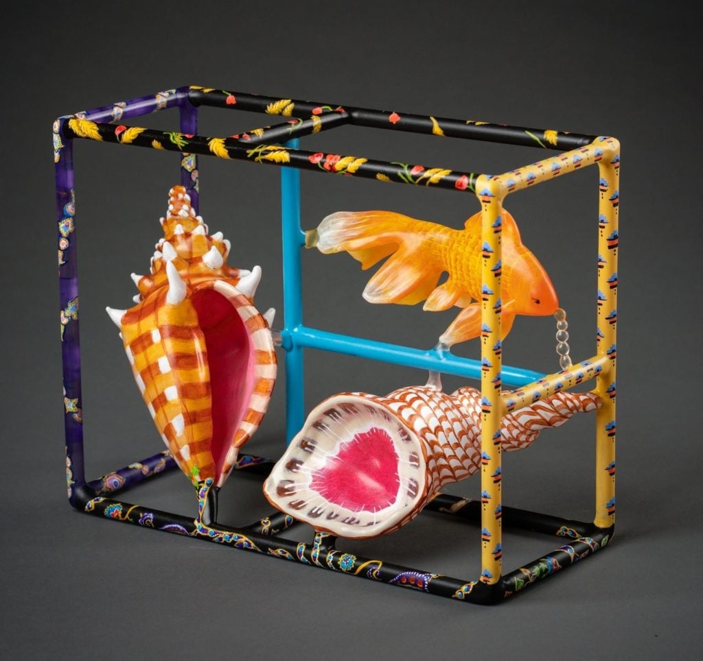A colorful glass box framework, with a glass goldfish and two glass shells suspended inside it.