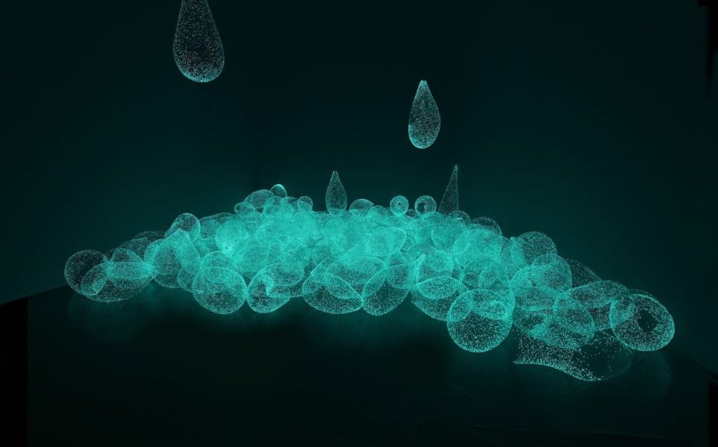 Three-dimensional glass teardrops lay in a pile in the corner of an otherwise empty and dark room, with two suspended from the ceiling as if falling. They are glowing a blue-green phosphorescent color, and each has small bubbles suspended in its surface.