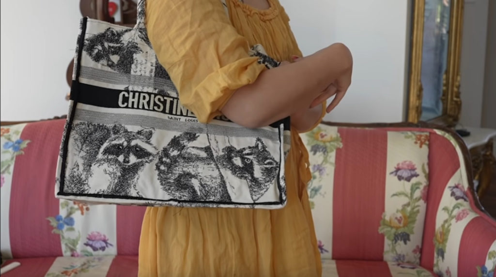 A woman in a yellow dress standing with a black and white tote bag slung over her shoulder. The tote bag is embroidered with detailed images of raccoons, as well as the name ‘Christine.’