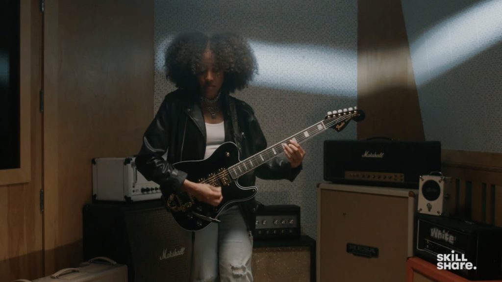 Guitarist Taylor Gamble stands while playing a guitar inside a sound booth. Behind her are amps and other recording equipment.