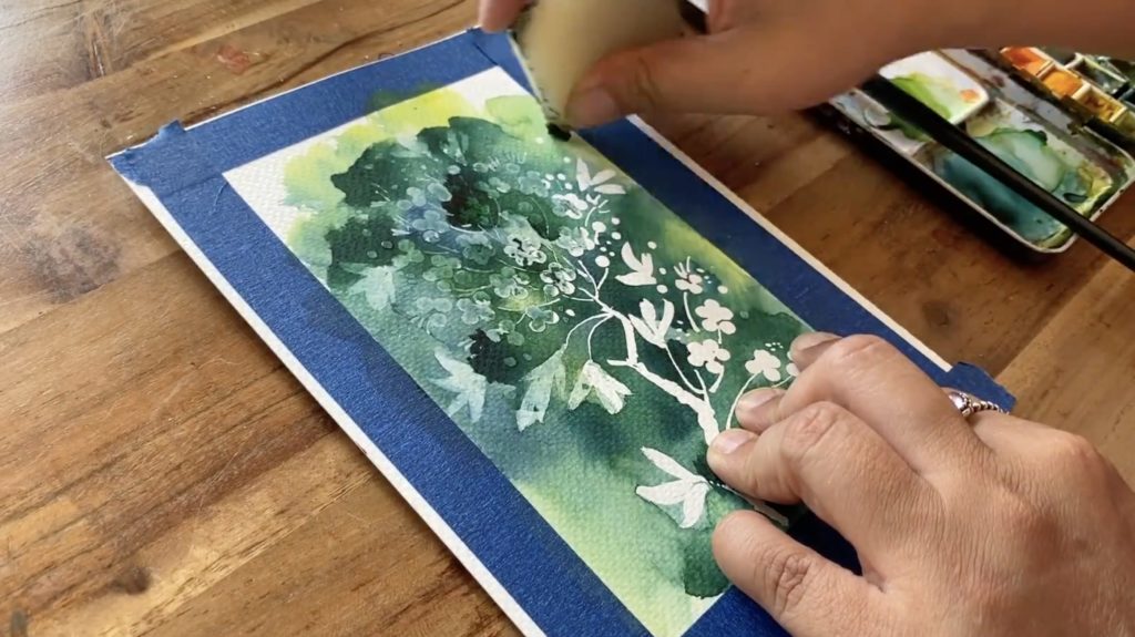 A watercolor painting of flowers and leaves, done in various shades of blue and green, with some of the flowers represented by negative space. The artist’s hands, watercolor palette, and paintbrush are also partially in the shot.
