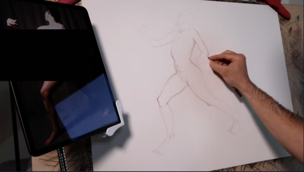 A nude figure drawing in mid-process. The artist's hand is visible in frame, shading an arm of the figure, and the reference illustration is also visible on the right hand side.