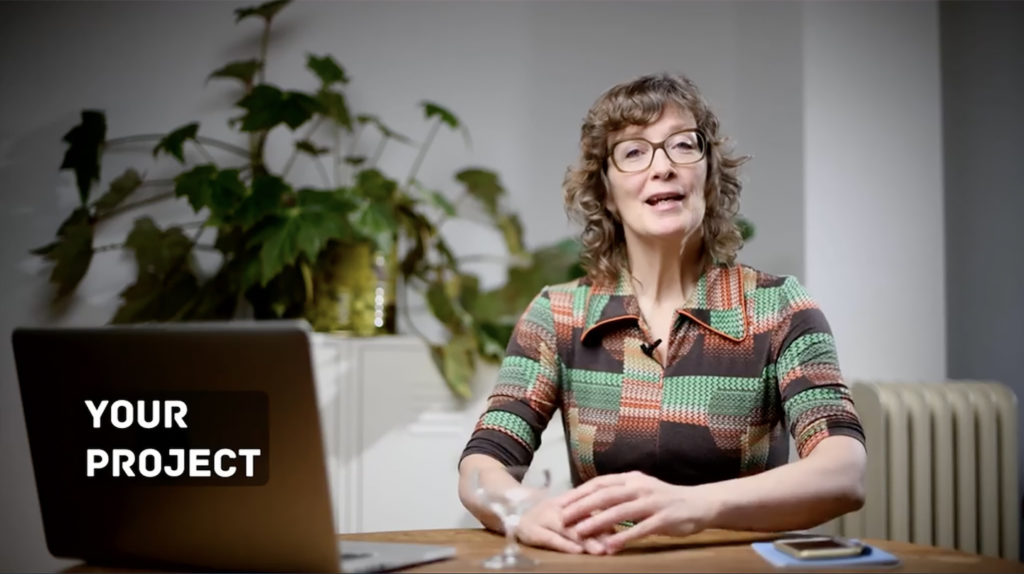 Skillshare teacher, Lucy Lambriex, sitting behind a desk with an open laptop on it. There's a large plant in the background. She's speaking to the camera.