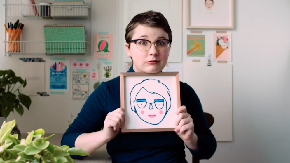 Photo of a woman with short brown hair and glasses holding up a picture frame under her chin. The picture frame holds a drawing of the woman, making the same facial expression.