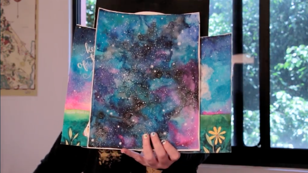 A hand holds a few watercolor paintings at the camera. The paintings are stacked on top of each other and area ll variations of night sky watercolors.