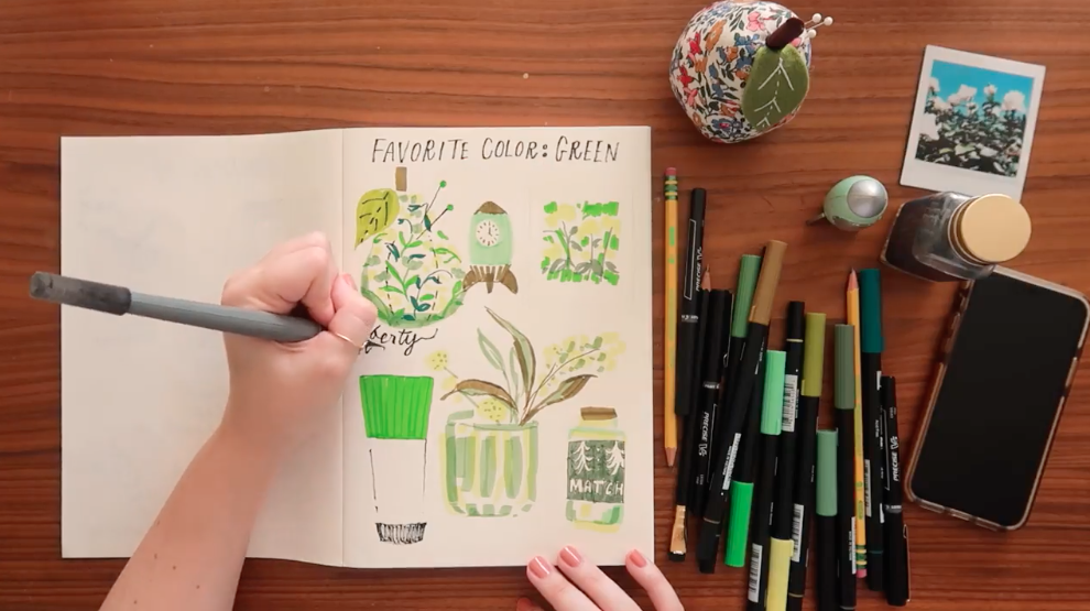 Overhead photo of an open sketchbook featuring the text "Favorite Color: Green" and drawings of various green objects. A hand holds a pen, and draws on the sketchbook. To the right of the frame is a pile of pens and markers in various shades of green, along with a polaroid picture, ink bottle, pear-shaped painted paper weight, and cell phone.