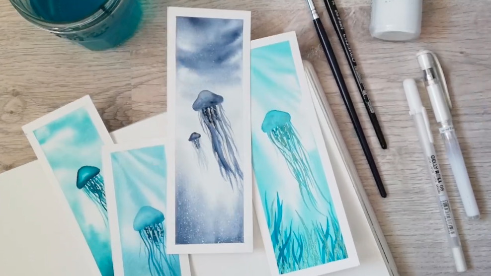 Overhead photo of 4 bookmarks with jellyfish painted in watercolors. To the right of the bookmarks are two paintbrushes, and two white pens.
