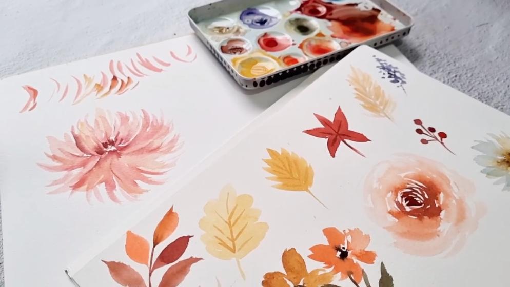 Two pieces of paper with watercolor sketches of various Autumn leaves and flower blossoms. A paint tray of watercolors sits at the top of the frame.