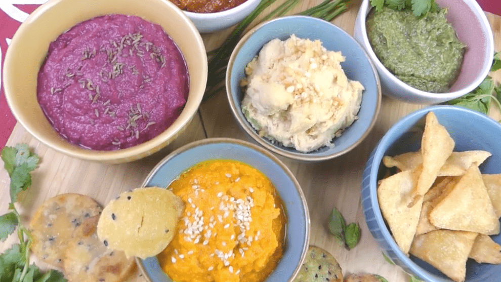 Pink, orange, green and cream-colored dips in brightly colored bowls, on a table with chips for dipping.