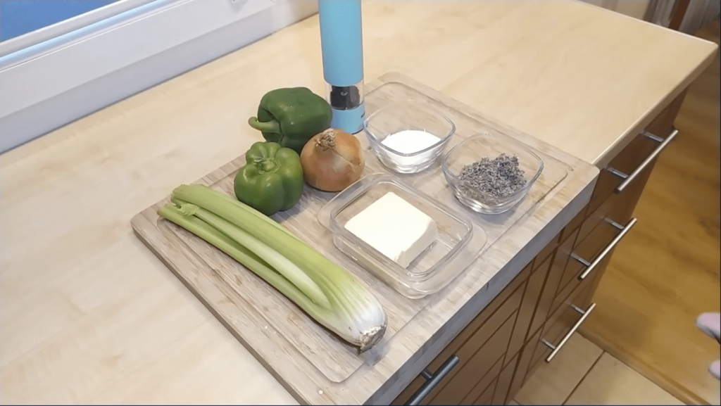Turkey stuffing ingredients sit on a cutting board which lays on a kitchen counter. The ingredients include celery, peppers, onion, salt, pepper, sage, and butter.