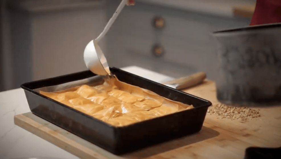 Syrup is carefully poured over baked baklava with a ladle.