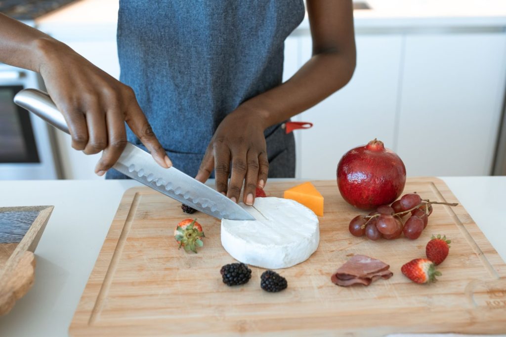 Using a shiny, silver knife, two hands cut into a hunk of round cheese on a wooden cutting board. Surrounding the cheese are a variety of fruits, meats, and cheeses, like a pomegranate and blackberries.