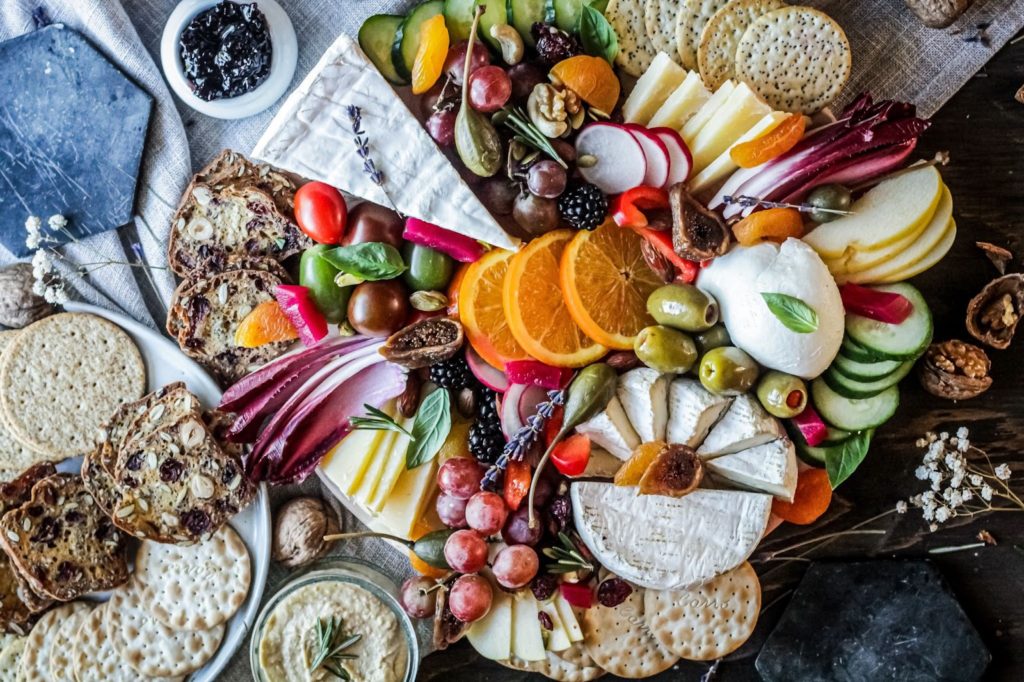 Three rings of oranges are surrounded by a variety of cheeses, fruits, vegetables, and crackers. With crispy cucumbers, deep-hued cabbage, and slices of creamy cheese, the board is a pop of color against a more neutral colored background.