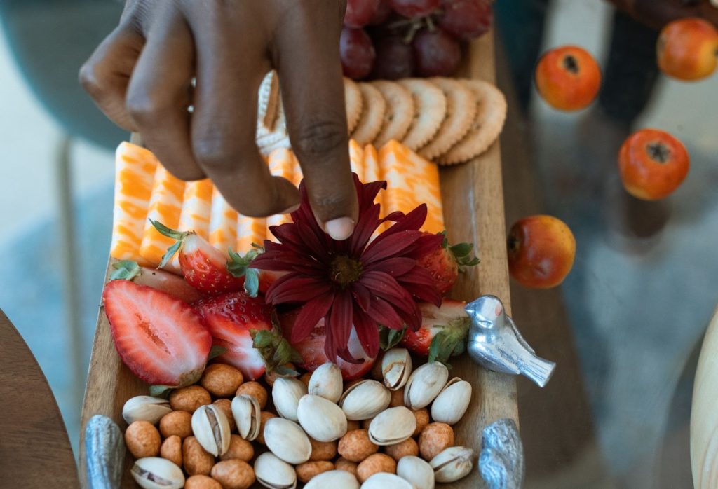 A hand places a burgundy flower onto a charcuterie board filled with cut strawberries, nuts, cheese, and crackers.