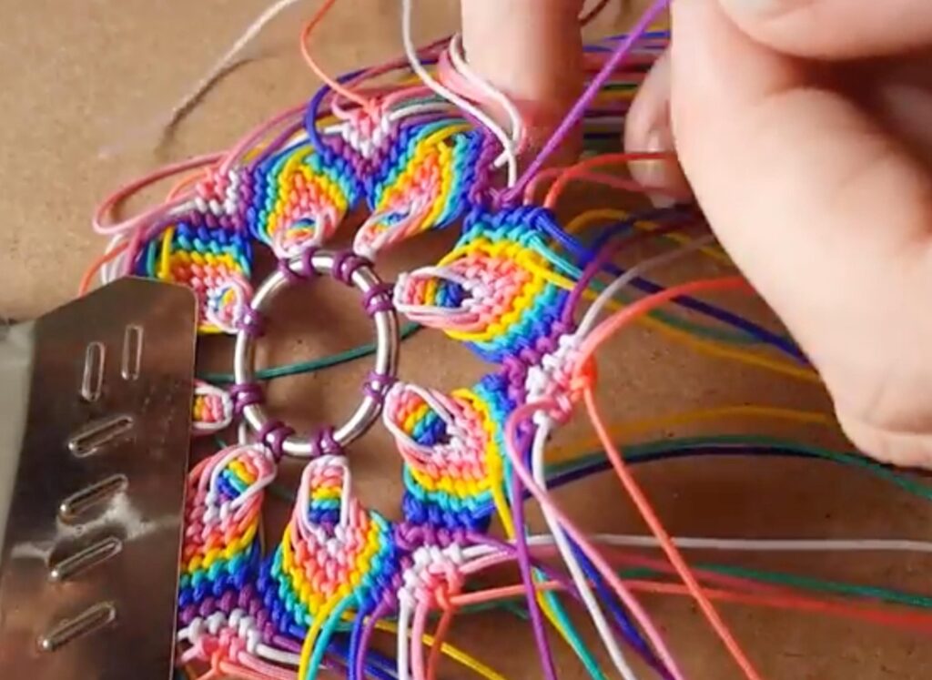 A close up of a macramé mandala with seven branches and seven colors of thread. The teacher has nearly finished making it and is tying together the final few knots. The unused thread is overhanging and the mandala is held in place by a clipboard clip. 