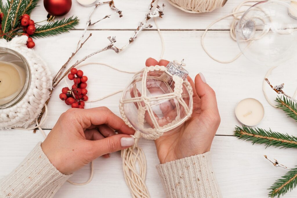 A woman holds a transparent Christmas bauble covered in a macramé pattern. The table is covered with festive touches, including pine needles, holly berries and a macramé candle cover.