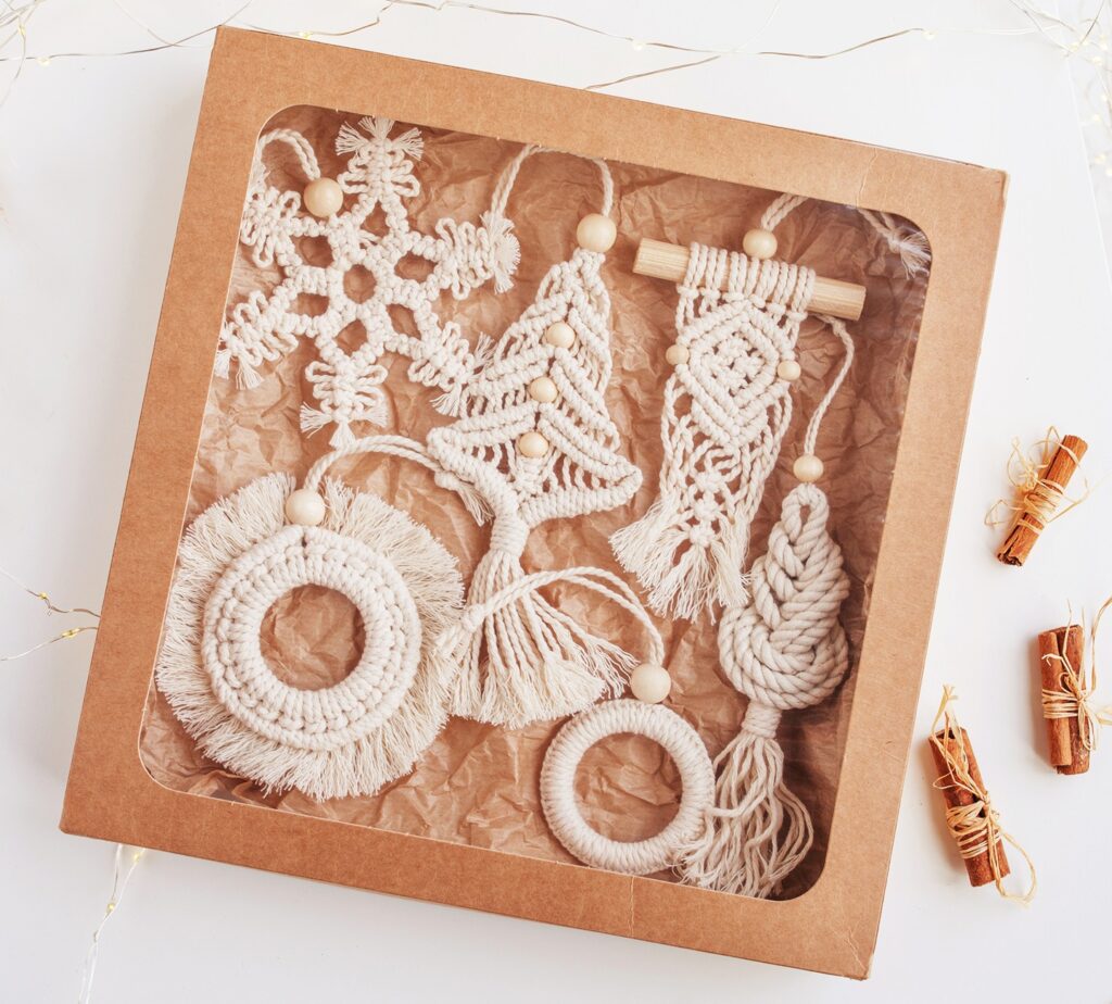 A group of macramé Christmas ornaments made from white cord, presented in a see-through box. The ornaments include a snowflake, a christmas tree, and other shapes to hang from the Christmas tree.