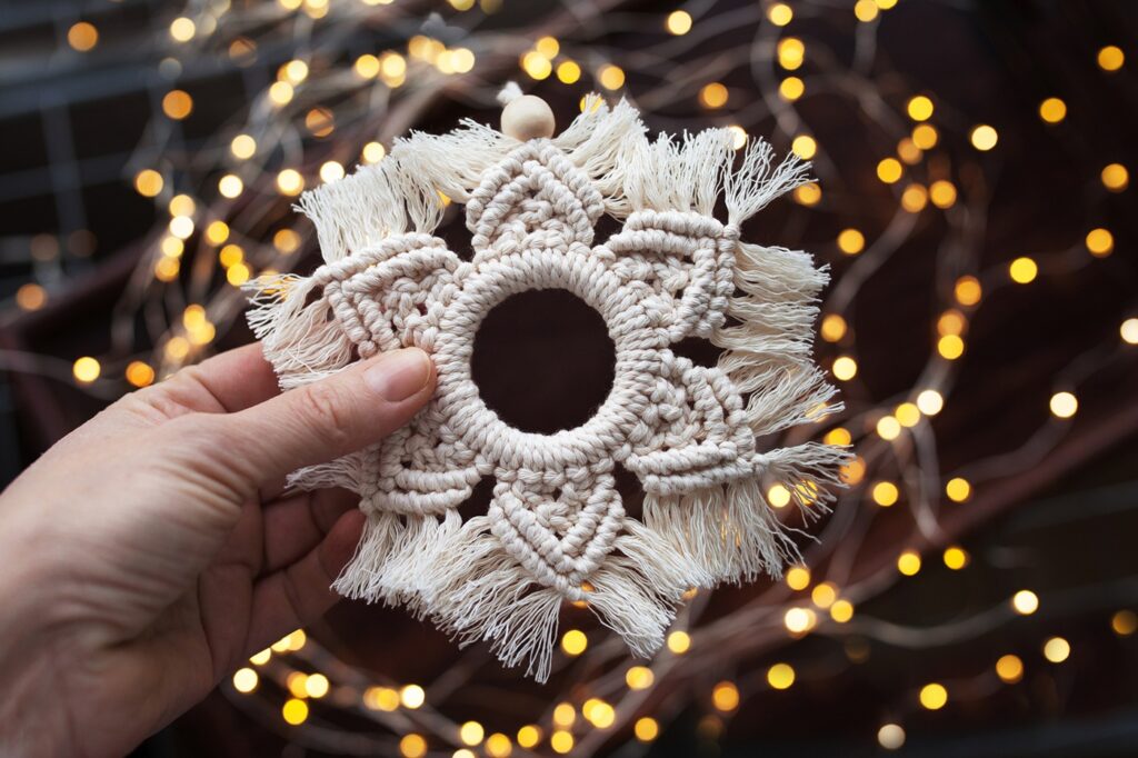 A hand holding a macramé snowflake ornament, with Christmas lights in the background. The snowflake is built around a central ring and has six branches. The excess cord has been combed to create a fringe effect. 