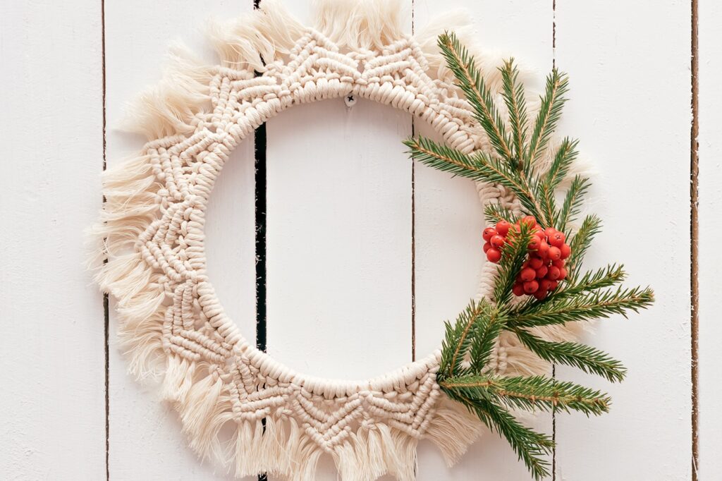 A macramé Christmas wreath made from a mandala wall hanging pattern. It uses white cord and has branches of pine and holly berries attached to feel more festive.